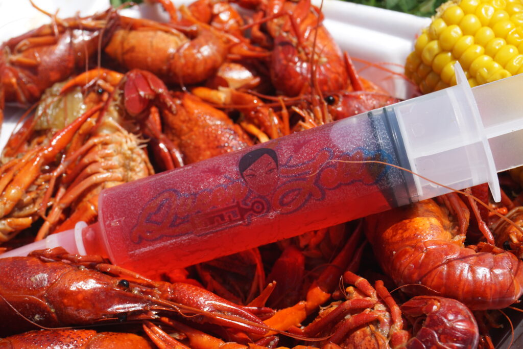 This image features a close-up view of a traditional crawfish boil, a staple at social gatherings in areas like Louisiana. In the foreground, a novelty cocktail syringe, branded with a cursive logo, rests atop the pile of freshly boiled crawfish, adding a unique and modern twist to the scene. Accompanying the spicy red crawfish is a piece of corn on the cob, rounding out the classic Southern meal. The syringe is filled with a crimson-colored liquid, suggesting a potent cocktail mix that complements the bold flavors of the seafood feast. This setting is often associated with communal dining and festive outdoor events where such cocktails might be enjoyed.