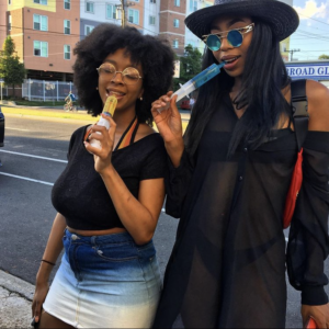 girl with afro smiles while slurping yellow jello shots and girl with hat and sunglasses slurps blue jello shots in syringe
