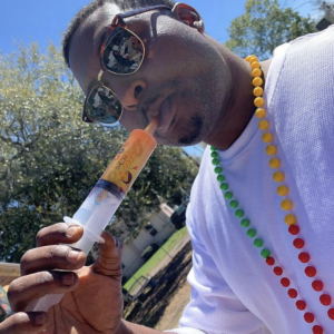 An individual wearing sunglasses and a white T-shirt is enjoying a sunny day outdoors. They are blowing into a novelty drink dispenser designed to resemble a cocktail push pop, with a label that includes the text "Queen Jelly." The person is also adorned with a colorful beaded necklace in a pattern of yellow, green, and red beads, evoking a festive atmosphere commonly associated with street parties or celebrations like Mardi Gras. The beads and the novelty cocktail dispenser suggest a playful and lighthearted mood, perfect for social gatherings or parades.