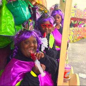 A trio of individuals are dressed in vibrant Mardi Gras costumes, featuring purple wigs and neon pink and green attire. They're seen enjoying the festivities, with two of them playfully using novelty cocktail syringes, hinting at the lively spirit of the event. Their accessories, including bejeweled tiaras and quirky glasses, add to the celebratory flair. The background shows a float adorned with bold, colorful designs, contributing to the overall atmosphere of a joyous parade. This image captures the essence of Mardi Gras, with its focus on fun, costumes, and unique ways to enjoy a cocktail.