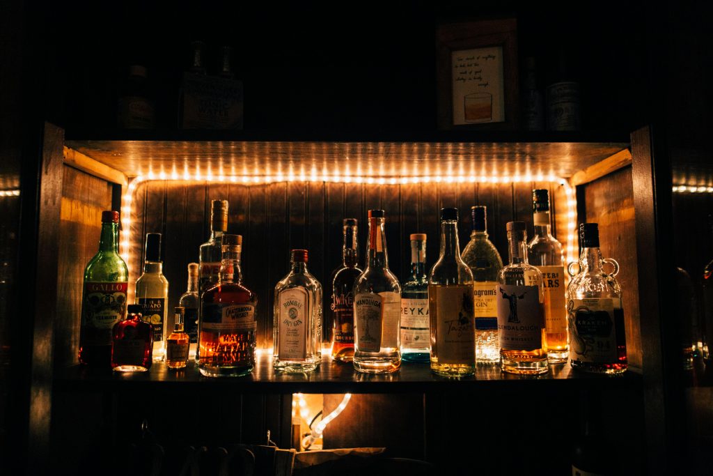 The photo shows a dimly lit wooden shelf displaying a variety of liquor bottles in a bar setting. The bottles, with labels facing forward, include different brands and types of spirits like gin, whiskey, and vermouth. Above the bottles, string lights provide a warm, ambient glow that highlights the bottles and casts a cozy atmosphere. There are a few framed items on the wall behind the shelf, adding to the decorative feel of the bar. The overall mood is inviting, reminiscent of an intimate speakeasy or a personal home bar.