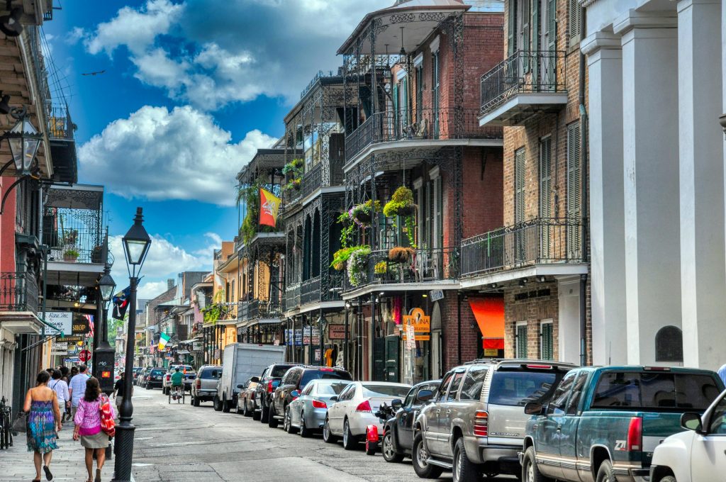 This image depicts a lively urban street scene, likely taken during a sunny day. The architecture suggests a historical area, with multi-storied buildings featuring ornate iron balconies adorned with hanging plants. The street is bustling with activity; pedestrians are walking on the sidewalks, and the road is lined with parked cars, suggesting a popular and possibly touristic neighborhood. The presence of flags and shop signs indicates commercial activity. The sky is partly cloudy, adding a dynamic backdrop to the urban landscape.
