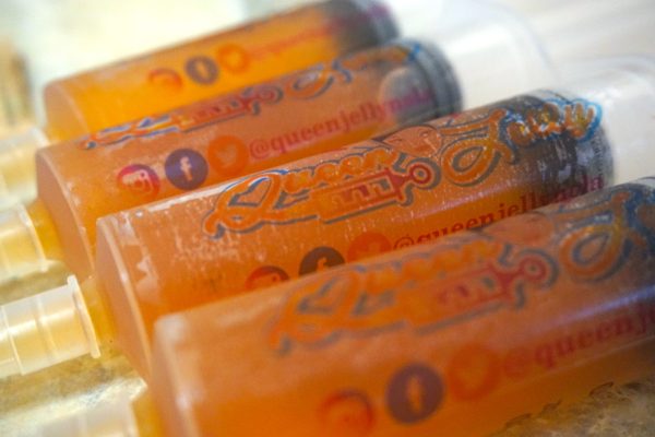 Close-up of Queen Jelly-branded syringes filled with vibrant, orange-colored gelatin, lined up in a row. The syringes are partially frosted, indicating they are chilled and ready to be enjoyed as jello shots. The Queen Jelly logo is prominently displayed on each syringe, along with social media icons in the background. Showing syringes for jello shot recipe